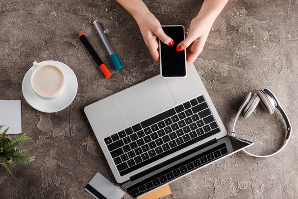 top view of woman holding smartphone near laptop, cup of coffee, plant, headphones and credit cards, e-commerce concept - Photo, Image