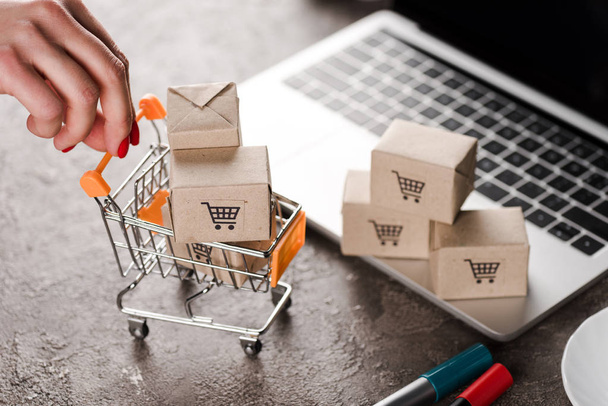 cropped view of woman holding toy shopping cart with small boxes near laptop, e-commerce concept - Photo, Image