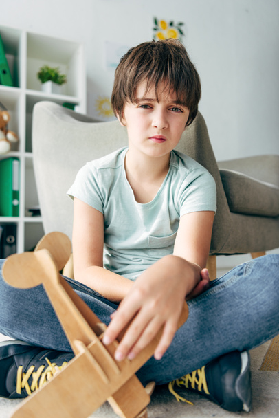 sad kid with dyslexia sitting on floor and holding wooden plane  - Photo, Image