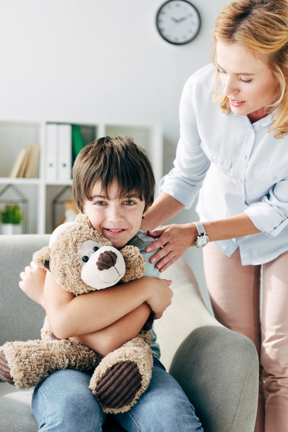 smiling kid with dyslexia holding teddy bear and child psychologist looking at him  - Φωτογραφία, εικόνα