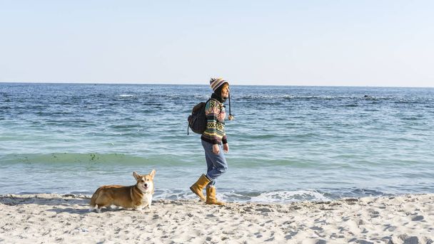 Young happy female walk with cute corgi dog on the autumn sunny beach - Fotoğraf, Görsel