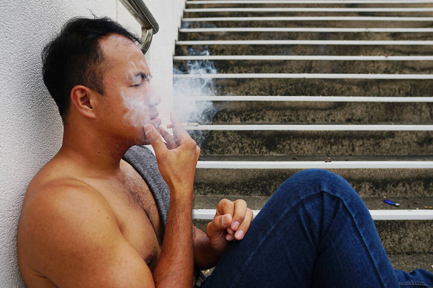 the sad depressed man without a t-shirt sitting on the stairs and smoking  - Photo, image