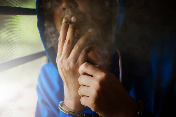 dark photo with the man with handcuffs in blue hoodie smoking near the wall, prisoner smoking with stress - Zdjęcie, obraz