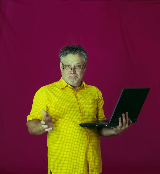Starší muž s šedivými vlasy drží v rukou notebook. V obličeji se projevují různé emoce. Radost, rozkoš, nejasnost, zamyšlení, rozkoš, něha. Portrét muže ve žluté košili na červeném pozadí s počítačem. - Fotografie, Obrázek