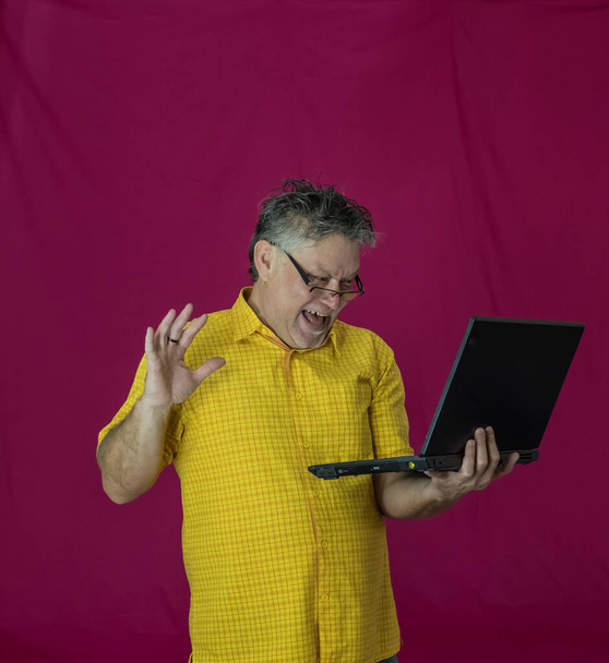 Ein älterer Mann mit grauen Haaren hält einen Laptop in der Hand. Verschiedene Emotionen kommen im Gesicht zum Ausdruck. Freude, Vergnügen, Dunkelheit, Nachdenklichkeit, Freude, Zärtlichkeit. Porträt eines Mannes in gelbem Hemd auf rotem Hintergrund mit Computer. - Foto, Bild
