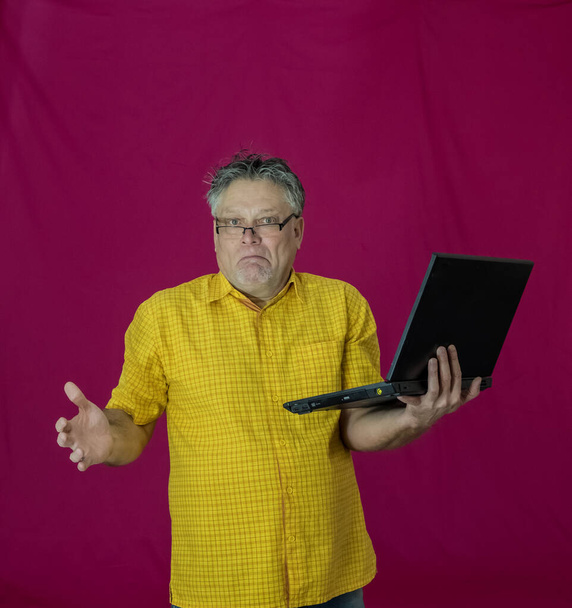 An elderly man with gray hair holds a laptop in his hands. Different emotions are expressed on the face. Joy, pleasure, obscurity, thoughtfulness, delight, tenderness. Portrait of a man in a yellow shirt on a red background with a computer. - Photo, Image