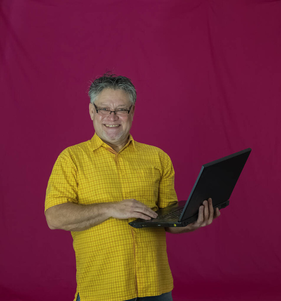Egy ősz hajú idős férfi laptopot tart a kezében. Az arcon különböző érzelmek jelennek meg. Öröm, öröm, homály, figyelmesség, öröm, gyengédség. Egy sárga inges, piros hátterű férfi portréja számítógéppel. - Fotó, kép