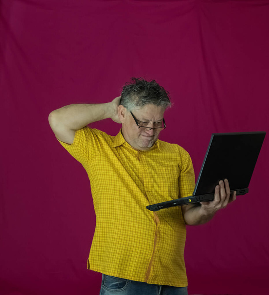 Um homem idoso com cabelo grisalho tem um portátil nas mãos. Emoções diferentes são expressas no rosto. Alegria, prazer, obscuridade, consideração, deleite, ternura. Retrato de um homem com uma camisa amarela em um fundo vermelho com um computador
. - Foto, Imagem