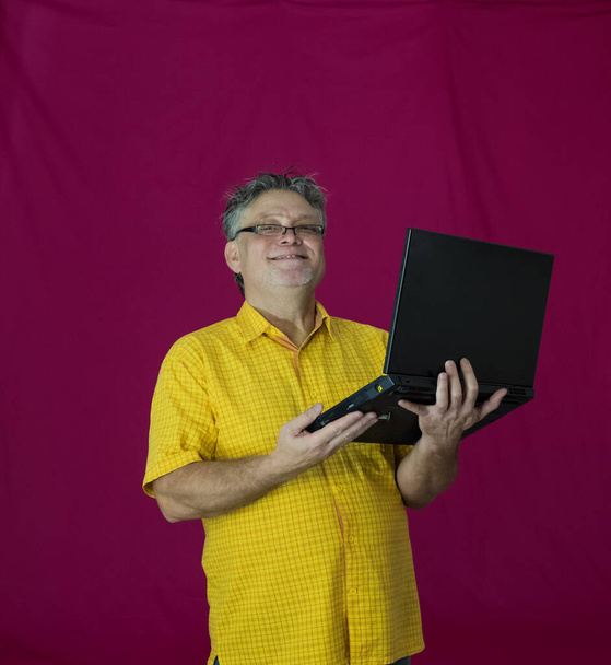 Starszy mężczyzna z siwymi włosami trzyma laptopa w rękach. Różne emocje wyrażane są na twarzy. Radość, przyjemność, ciemność, troskliwość, rozkosz, czułość. Portret mężczyzny w żółtej koszuli na czerwonym tle z komputerem. - Zdjęcie, obraz