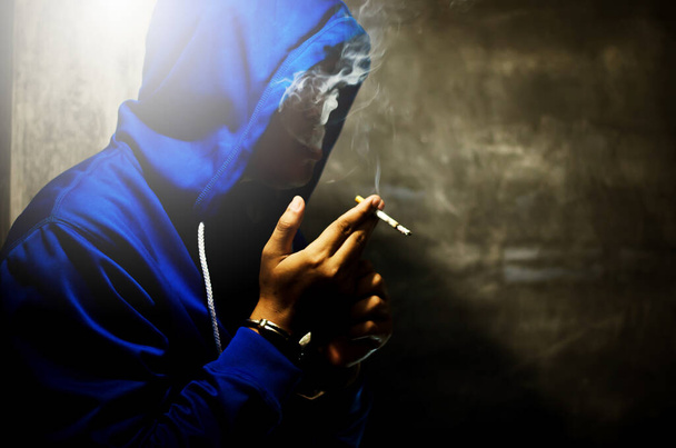 dark photo with the man in blue hoodie smoking near the wall in stage light  - Φωτογραφία, εικόνα