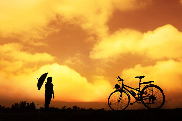 silhouette of the unrecognizable girl with bicycle and umbrella at sunset with beautiful clouds and horizon   - Фото, зображення