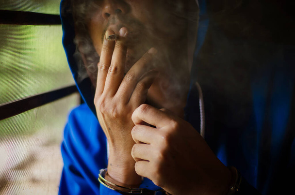 dark photo with the man with handcuffs in blue hoodie smoking near the wall, prisoner smoking with stress - Φωτογραφία, εικόνα
