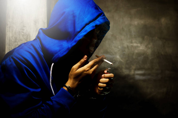 dark photo with the man in blue hoodie smoking near the wall in stage light  - Zdjęcie, obraz