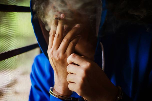dark photo with the man with handcuffs in blue hoodie smoking near the wall, prisoner smoking with stress - Zdjęcie, obraz