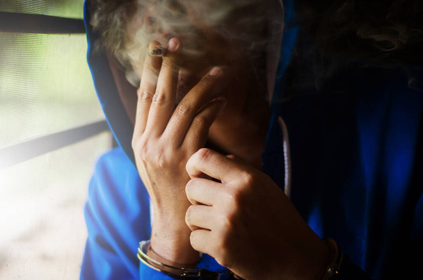 dark photo with the man with handcuffs in blue hoodie smoking near the wall, prisoner smoking with stress - Фото, изображение