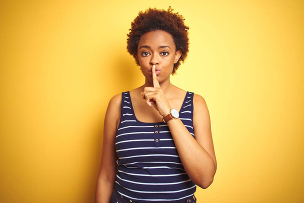 Beauitul afro-amerikai nő nyári pólót visel izolált sárga háttér kérve, hogy maradjon csendben ujjal a száján. Csend és titkos koncepció. - Fotó, kép