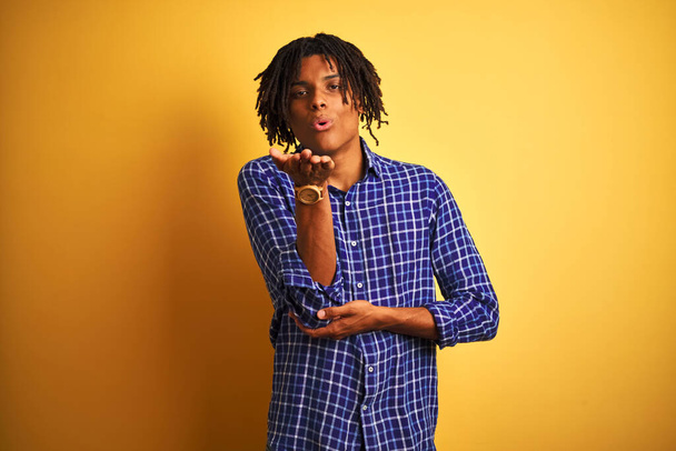Afro man with dreadlocks wearing casual shirt standing over isolated yellow background looking at the camera blowing a kiss with hand on air being lovely and sexy. Love expression. - Photo, Image