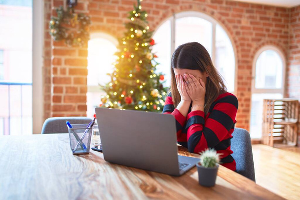 Beautiful woman sitting at the table working with laptop at home around christmas tree with sad expression covering face with hands while crying. Depression concept. - Φωτογραφία, εικόνα