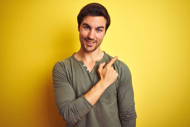 Jonge knappe man draagt casual t-shirt staan over geïsoleerde gele achtergrond vrolijk met een glimlach van het gezicht wijzen met hand en vinger naar de zijkant met een vrolijke en natuurlijke uitdrukking op het gezicht - Foto, afbeelding