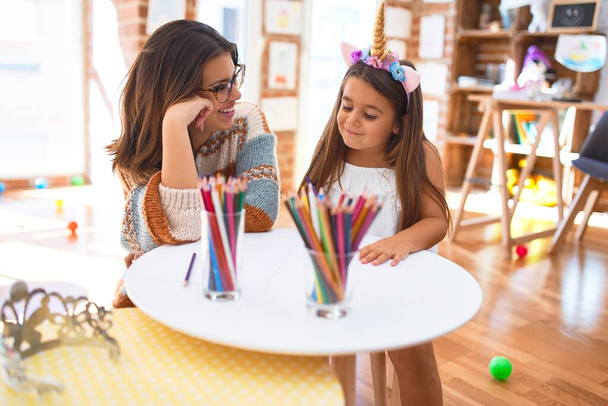 Bela professora e criança vestindo unicórnio desenho diadema usando papel e lápis em torno de lotes de brinquedos no jardim de infância
 - Foto, Imagem