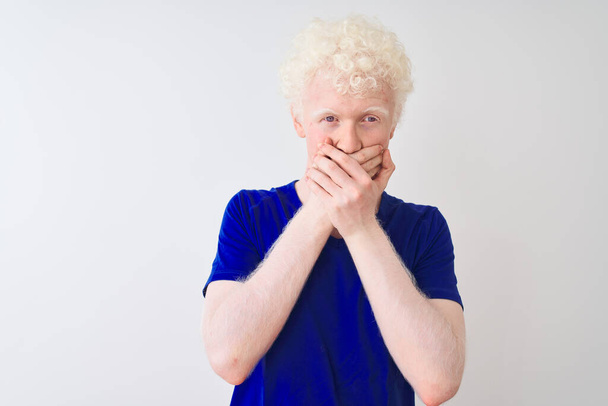 junger Albino-blonder Mann in blauem lässigem T-Shirt, der vor isoliertem weißem Hintergrund steht, schockiert, als er fälschlicherweise den Mund mit Händen bedeckt. Geheimes Konzept. - Foto, Bild