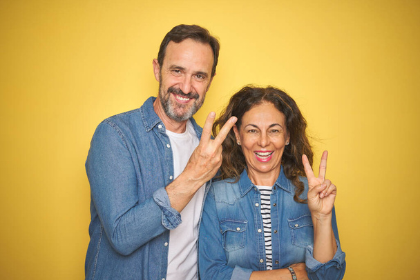 Bella coppia di mezza età insieme indossando camicia di jeans su sfondo giallo isolato sorridente con volto felice ammiccando alla fotocamera facendo segno di vittoria. Numero due.
. - Foto, immagini