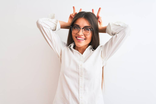 Молодая красивая деловая женщина в очках, стоящих на изолированном белом фоне, изображая смешного и сумасшедшего с пальцами на голове, как кроличьи уши, улыбаясь весело
 - Фото, изображение