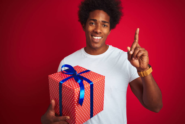 Молодой африканский американец держит подарок на день рождения стоя на изолированном красном фоне удивленный идеей или вопросом указывая пальцем со счастливым лицом, номер один
 - Фото, изображение