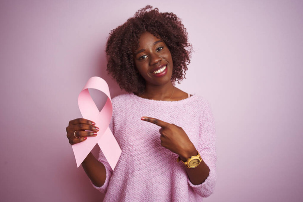 Young african afro woman holding cancer ribbon standing over isolated pink background very happy pointing with hand and finger - Zdjęcie, obraz