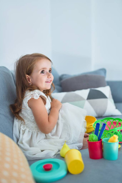Urocze blond maluch siedzi na kanapie gotowanie z jedzeniem i sztućce zabawki w domu - Zdjęcie, obraz