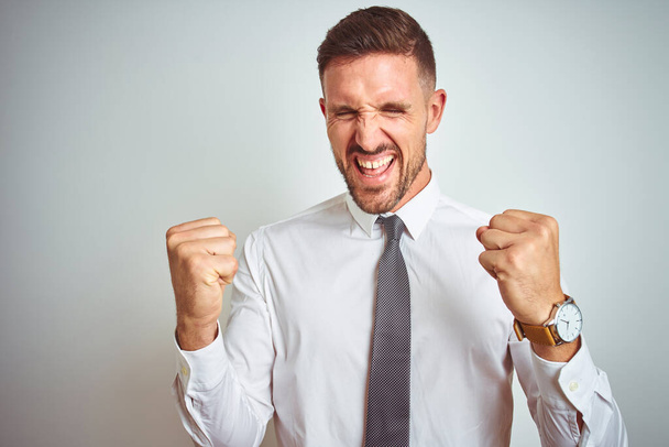 Young handsome business man wearing elegant white shirt over isolated background very happy and excited doing winner gesture with arms raised, smiling and screaming for success. Celebration concept. - Fotografie, Obrázek