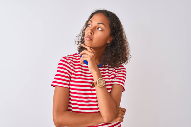 若いブラジル人女性が赤い縞模様のTシャツを着て、あごの質問について考えて手で孤立した白い背景の上に立って、集中的な表現。思慮深い顔で微笑んだ。疑いの概念. - 写真・画像