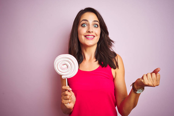 Young beautiful woman eating sweet candy over pink isolated background screaming proud and celebrating victory and success very excited, cheering emotion - Foto, afbeelding