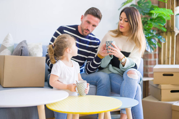 Beautiful family, parents sitting on the sofa drinking coffee looking his kid playing at new home around cardboard boxes - Foto, Imagen