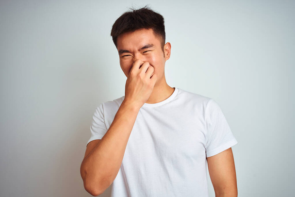 Jeune homme chinois asiatique portant t-shirt debout sur fond blanc isolé sentant quelque chose de puant et dégoûtant, odeur intolérable, retenant souffle avec les doigts sur le nez. Concept de mauvaises odeurs
. - Photo, image