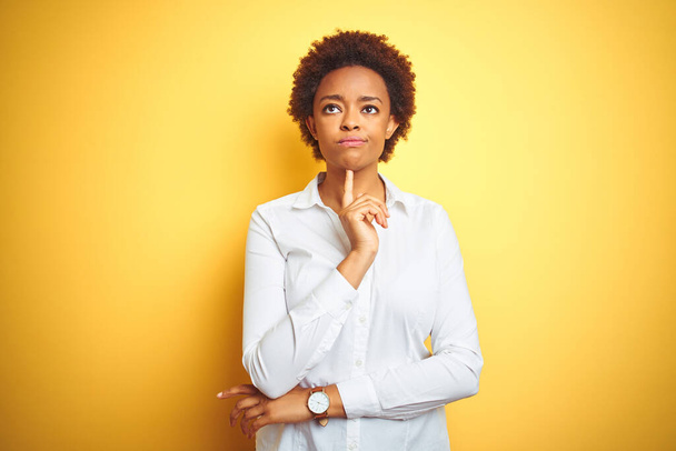 孤立した黄色の背景の上にアフリカ系アメリカ人のビジネス女性顎に指で疑いに集中し、疑問を見上げ考える - 写真・画像