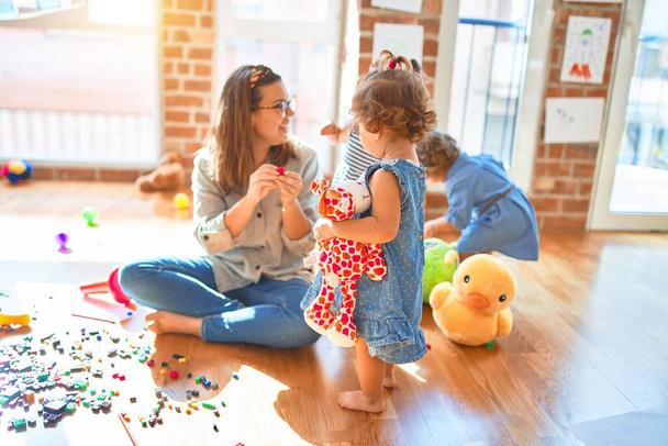 Hermosa maestra y grupo de niños pequeños jugando alrededor de muchos juguetes en el jardín de infantes
 - Foto, Imagen