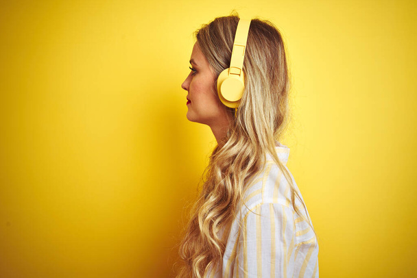 Νεαρή όμορφη γυναίκα ακούγοντας μουσική χρησιμοποιώντας ακουστικά πάνω από κίτρινο απομονωμένο φόντο κοιτάζοντας προς τα πλάγια, να χαλαρώσετε προφίλ ποζάρουν με φυσικό πρόσωπο με αυτοπεποίθηση χαμόγελο. - Φωτογραφία, εικόνα