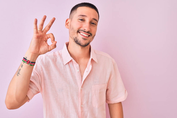 Jonge knappe man draagt roze shirt staan over geïsoleerde achtergrond lachend positief doen ok teken met de hand en vingers. Succesvolle uitdrukking. - Foto, afbeelding