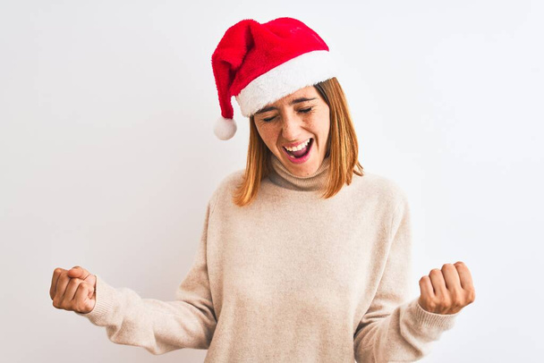 Bella donna rossa indossa cappello di Natale su sfondo isolato molto felice ed eccitato facendo gesto vincitore con le braccia alzate, sorridente e urlando per il successo. Concetto di celebrazione
. - Foto, immagini