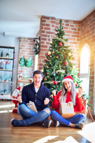 クリスマスツリーの周りの床に座っているサンタクラスの帽子をかぶっている若いカップルは、腕を上げて目を開いて成功したことに驚いて驚いて驚いています。勝者のコンセプト. - 写真・画像