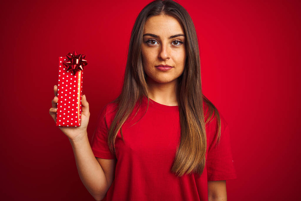 Νεαρή όμορφη γυναίκα κρατώντας δώρο γενεθλίων στέκεται πάνω από απομονωμένο κόκκινο φόντο με μια σίγουρη έκφραση στο έξυπνο πρόσωπο σκέψης σοβαρή - Φωτογραφία, εικόνα