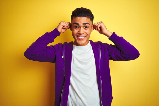 Młody Brazylijczyk ubrany w purpurową bluzę stojący nad odizolowanym żółtym tłem Uśmiechnięty pociągający za uszy palcami, zabawny gest. Problem przesłuchań - Zdjęcie, obraz