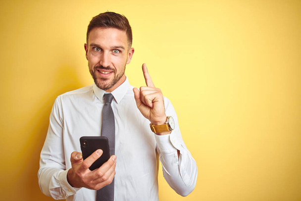 junger gutaussehender Geschäftsmann mit Smartphone über gelbem isolierten Hintergrund überrascht mit einer Idee oder Frage mit glücklichem Gesicht, Nummer eins - Foto, Bild