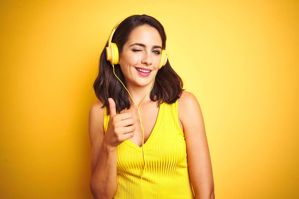 Young beautiful woman listening to music using headphones over yellow isolated background doing happy thumbs up gesture with hand. Approving expression looking at the camera with showing success. - Photo, Image