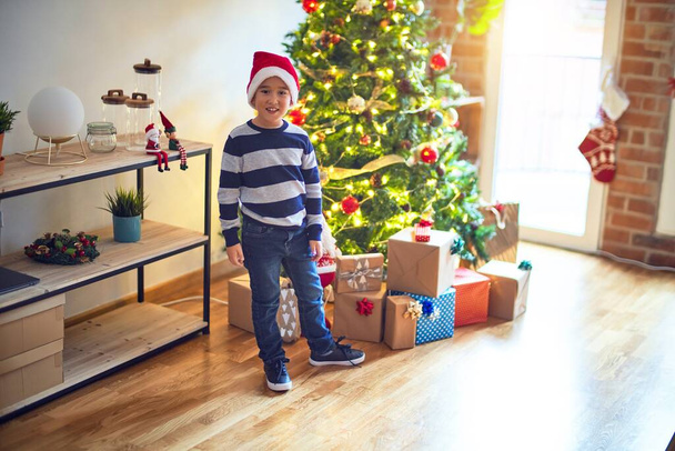 Suloinen lapsi hymyilee onnellisena ja itsevarmana. Seisoo yllään joulupukin hattu hymy kasvoillaan ympäri joulukuusi ja paljon lahjoja kotona
 - Valokuva, kuva