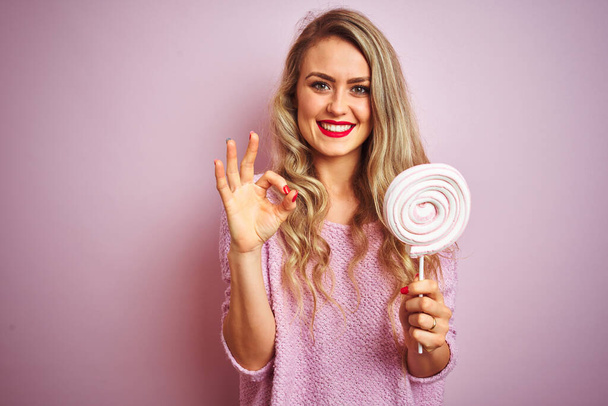 Mladá krásná žena pojídáním sladkých sladkostí na růžovém izolovaném pozadí dělá dobře nápis prsty, skvělý symbol - Fotografie, Obrázek