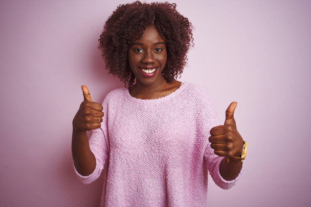 隔離されたピンクの背景の成功の印の上に立つセーターを身に着けている若いアフリカのアフロ女性は手で肯定的なジェスチャーをし、親指を笑顔と幸せ。陽気な表情と勝者のジェスチャー. - 写真・画像