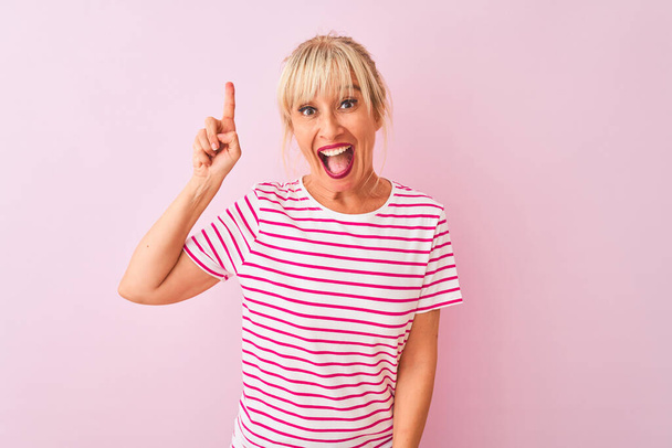 Femme d'âge moyen portant t-shirt rayé debout sur fond rose isolé pointant doigt vers le haut avec idée réussie. Sorti et heureux. Numéro un
. - Photo, image