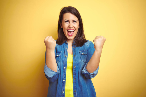 Young beautiful brunette woman wearing casual denim jacket over yellow isolated background celebrating surprised and amazed for success with arms raised and open eyes. Winner concept. - Photo, Image